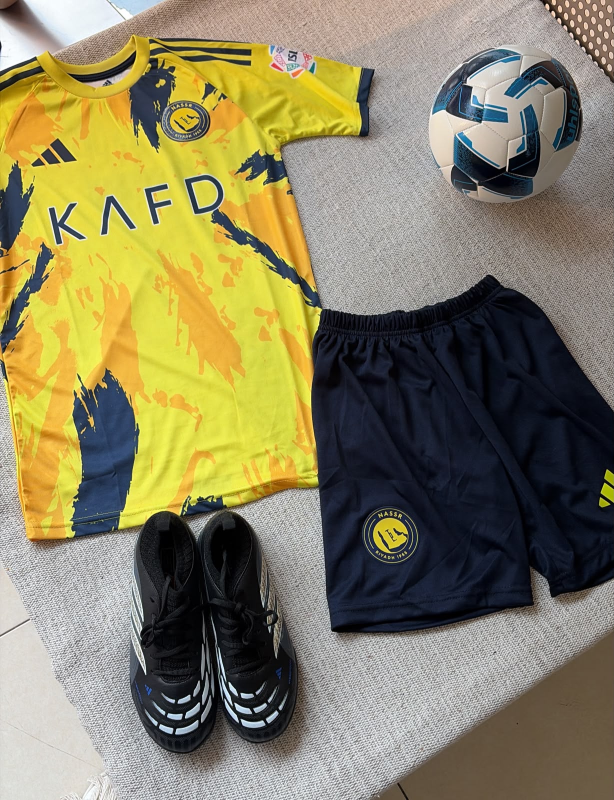 Al-Nassr kids football kit yellow KAFD jersey with navy brushstroke pattern and navy shorts flat lay