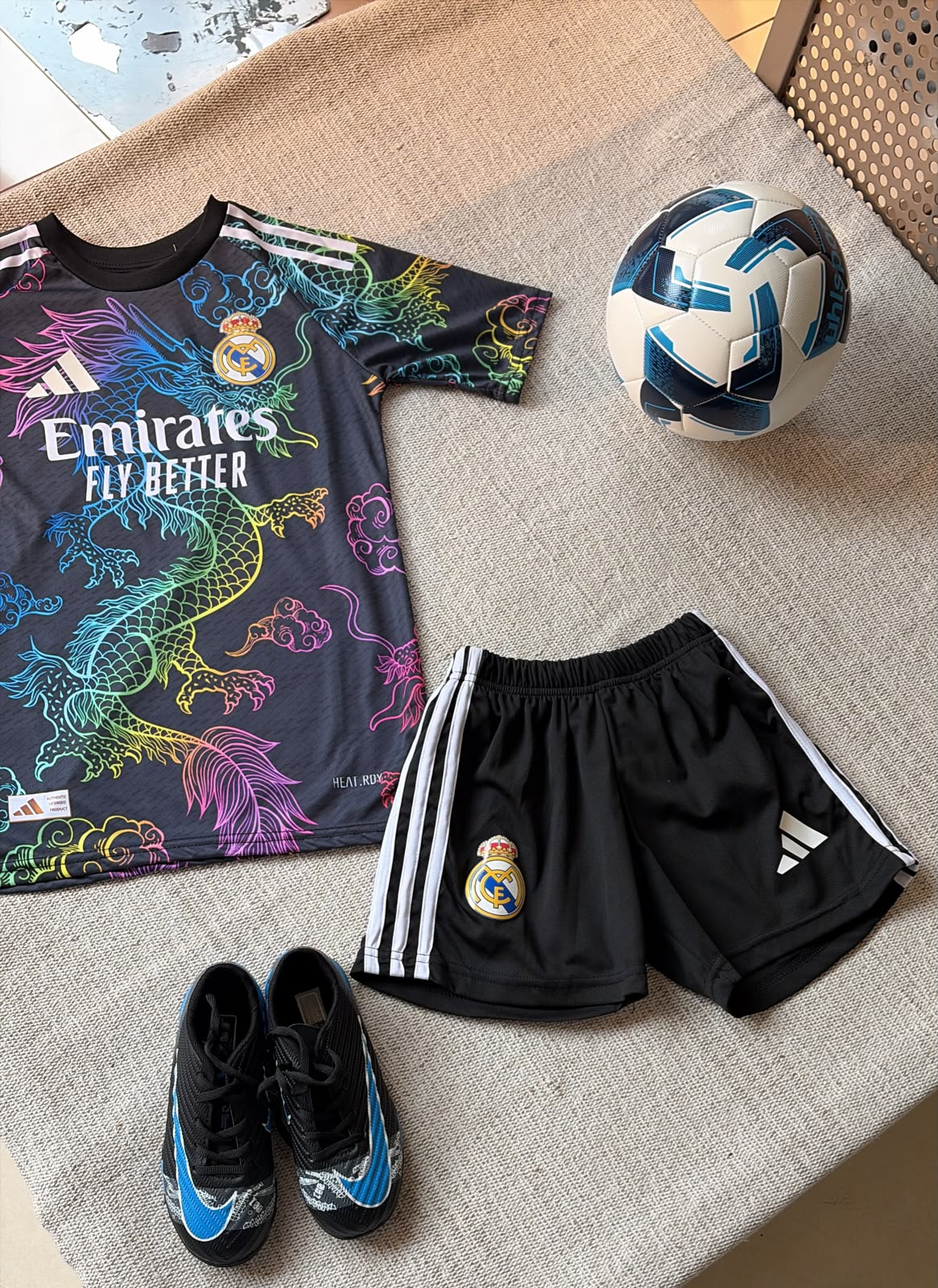 Real Madrid dragon kids football kit black jersey with neon dragon design and black Adidas shorts flat lay
