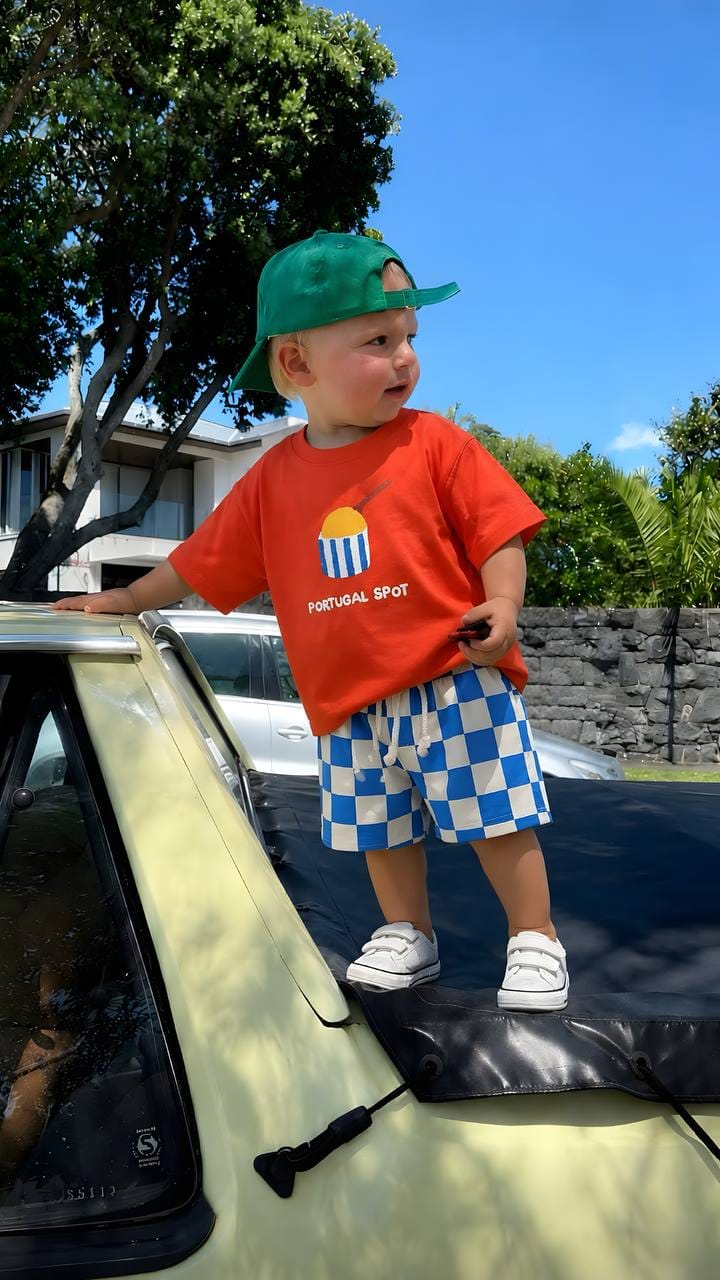 Baby wearing a red-orange Portugal Spot t-shirt and blue checkered shorts